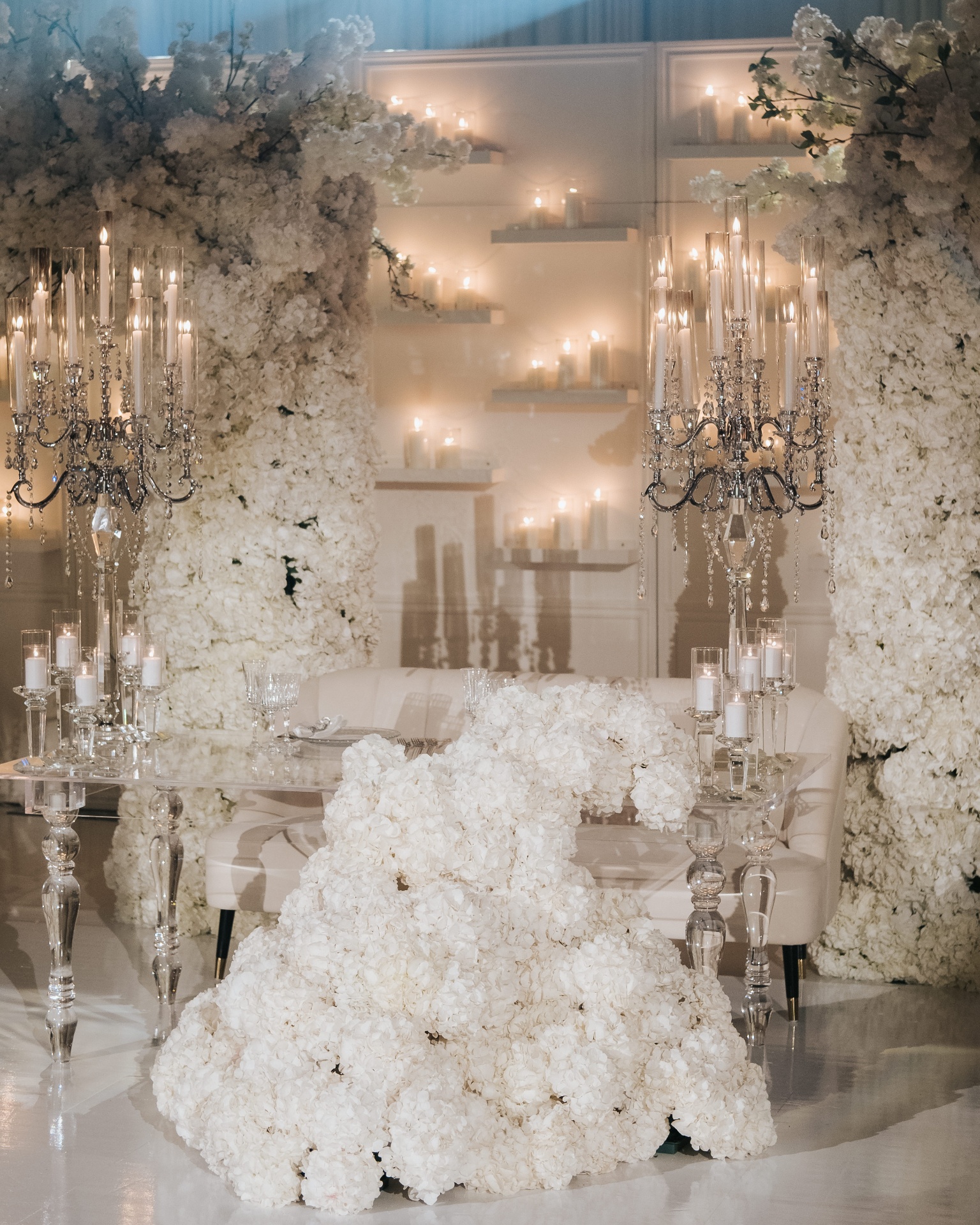 All-White Hydrangea Sweetheart - All-white hydrangea sweetheart table with towering crystal candelabras, cascading floral pillars, and candlelit votive wall by Aura Floral Design Miami
