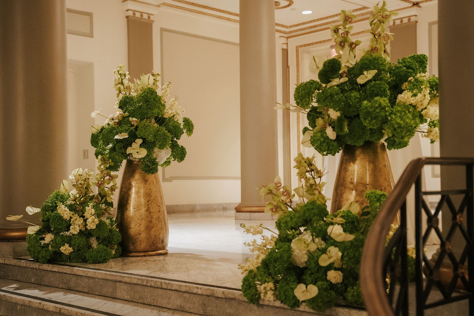 Grand Staircase Vases - Gold vessels overflowing with green and ivory blooms on a marble staircase by Aura Floral Design Miami